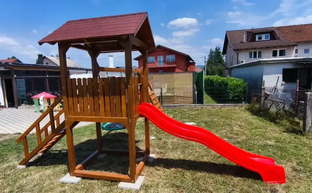 Sigurno drveno igralište za djecu s toboganom, smješteno u zelenom dvorištu pod vedrim nebom.