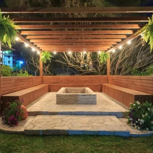 Vanjska pergola s rasvjetom, biljkama i klupama oko kamina, idealna za večernja druženja u vrtu