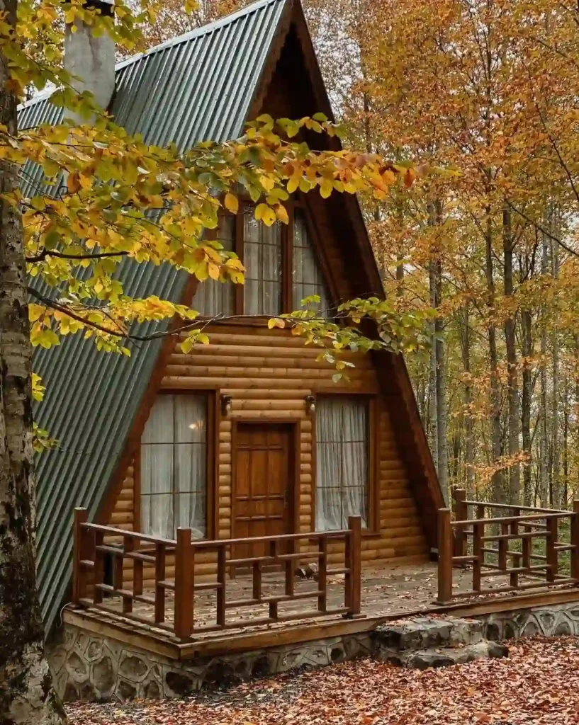 A-frame drvena kućica smještena u šumi tijekom jeseni, s kosim zelenim metalnim krovom koji se spušta gotovo do tla. Kućica ima malu drvenu terasu s ogradom, a ulazna vrata su smještena između dva velika prozora s zavjesama.