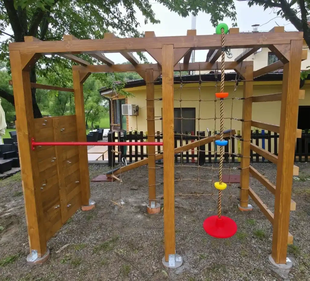 Drveni dječji poligon s penjalicom, konopcem, visećim diskovima i mrežastim zidom za penjanje na vanjskoj površini.